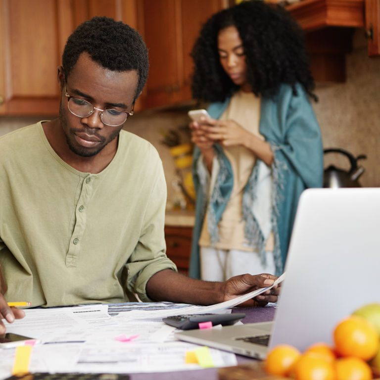 man sitting in kitchen looking at paper using a calculator with a woman on her mobile phone man sitting in kitchen looking at paper using a calculator with a woman on her mobile phone